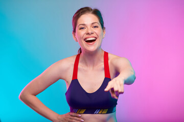 Fototapeta na wymiar Coach Girl points a finger straight in front of her. Happy young adult sporty woman in fitness bra sportswear. Female fitness portrait isolated on neon multicolor background.