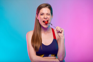 Sporty woman in fitness sportswear eating tomato on fork. Female fitness portrait isolated on neon multicolor background.