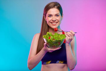 Smiling woman in sportswear eating green salad in glass bowl. Female fitness portrait isolated on neon multicolor background.