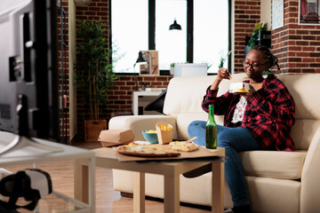 Female adult using chopsticks to eat noodles from delivery box, enjoying meal in front of television watching movie. Eating fast food takeout package as dinner, looking at film on tv.