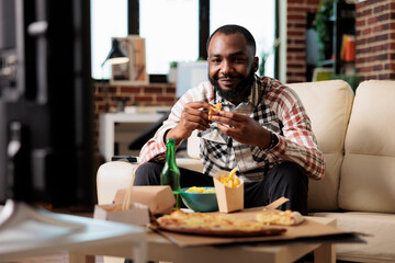 African american man eating slice of pizza from takeout delivery to watch television film in living...