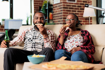 African american couple eating slice of pizza and drinking bottles of beer while they watch movie on tv together. Enjoying fast food takeaway delivery and beverage, watching television.