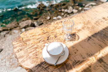 Cappuccino latte coffee cup mug and a glass of water standing on a wooden table in outside cafe with scenery sea and mountains in the background. Copy blank space.