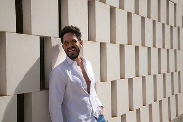 Handsome young man with beard, sunglasses and open white shirt, leaning against a wall of white...