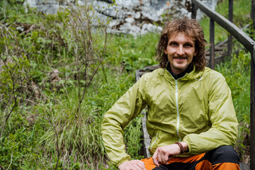 A cute guy smiles sitting in nature, a tourist on a trip looks into the camera, a portrait of a young man on a hike, curly hair of a hipster.