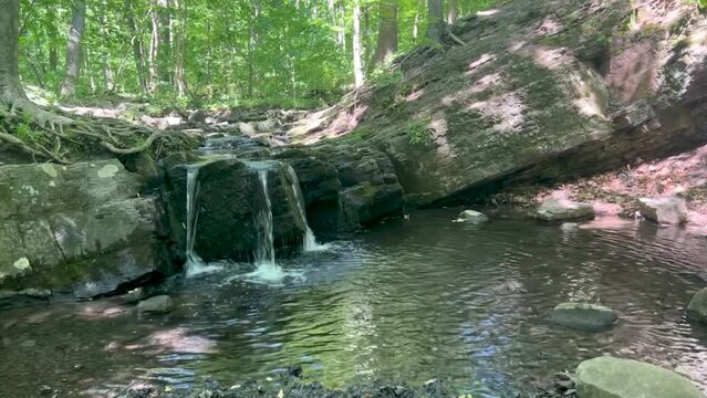 An idyllic enchanted woodland stream waterfall pours over a large rock formation with visible strata with green trees in the background reflected in the tranquil foreground pool serene nature with no 