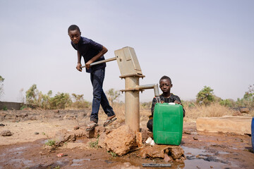 Two African boys busy filling water containers at a remote village pump; water scarcity in...