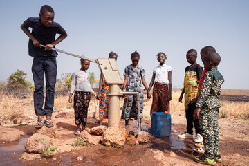 Several young African village children grouped around a manual pump filling canisters with clean and fresh drinking water