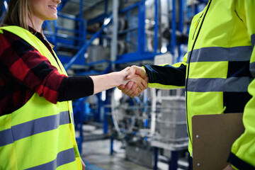 Engineer and industrial worker in uniform shaking hands in large metal factory hall and talking. Close-up.