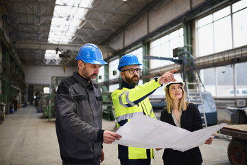Fototapeta na wymiar Manager supervisor, engineer and industrial worker in uniform discussing blueprints in large metal factory hall.