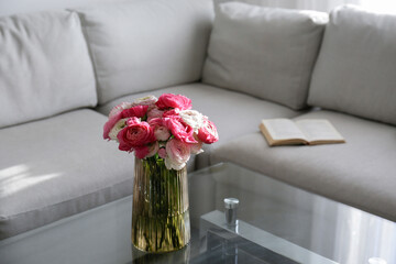 Close up shot of glass coffee table with bouquet of beautiful bicolor ranunculus flowers in a vase on foreground and gray textile couch on the background. Copy space, close up, natural light.