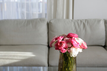 Close up shot of glass coffee table with bouquet of beautiful bicolor ranunculus flowers in a vase on foreground and gray textile couch on the background. Copy space, close up, natural light.