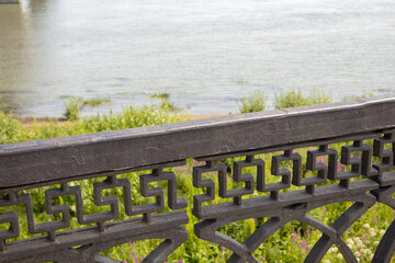 Obraz premium Engineering design of black cast-iron wrought-iron railings of the embankment fence, close-up against the background of the river and grass on the shore