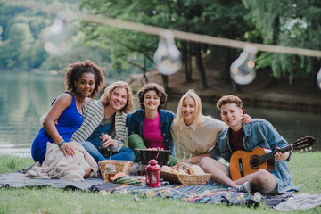 Group of friends having fun on picnic near a lake, sitting on blanket eating and playing guitar.