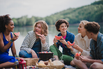 Group of young friends having fun on picnic near a lake, sitting on blanket and eating watermelon.