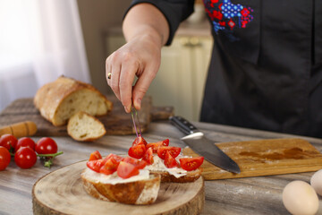 Female chef preparing breakfast in a bright country kitchen. He cuts the bread and spreads it with cheese. Kitchen table with flowers and kitchen board, bread, tomatoes, greens
