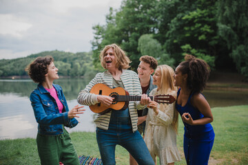 A group of young friends having fun on picnic near a lake, laughing and playing guitar.