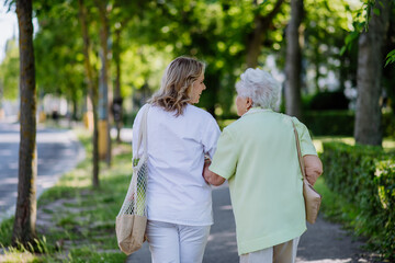 Rear view of caregiver with senior woman on walk in park with shopping bag.