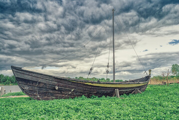 Old wooden boat in green grass near lake by dramatic dark blue sky. Summer landscape with ancient...