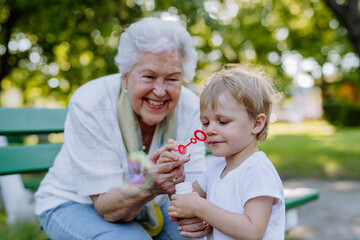 Great grandmother sitting on bench with her granddaughter and blowing soap bubbles together,...