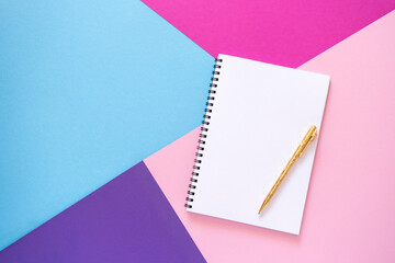 Empty notebook with gold pen lies on pink, purple, violet and light blue paper background....