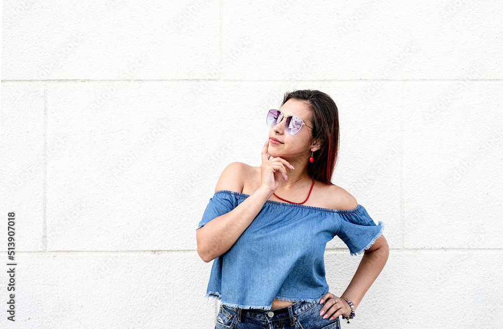 Wall mural stylish young woman wearing jeans shirt, sunglasses and bag, at street, white wall background