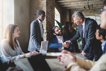Two businessmen make a deal by shaking hands sitting at a table together with other multiracial...