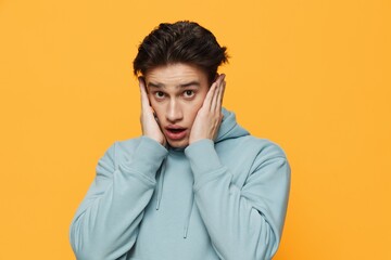 a close horizontal photo of a shocked young man standing on an orange background in a light blue hoodie, covering his ears with his hands, and looking very emotionally into the camera
