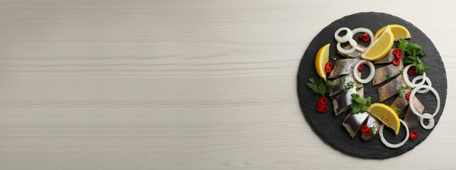 Fototapeta premium Slate plate with sliced salted herring fillet, parsley, chili pepper, onion rings and lemon on light beige wooden table, top view with space for text. Banner design