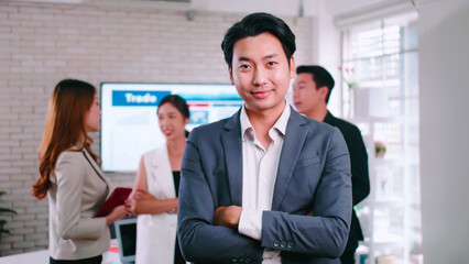 Confident young businessman standing with folded arms smiling at the camera in a boardroom with colleagues in the background.