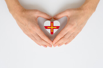 Obraz premium National flag of Guernsey in the shape of a heart in female hands on a light background.