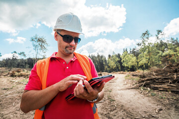 Forestry engineer wearing helmet holds digtal tablet in hands for quality material control