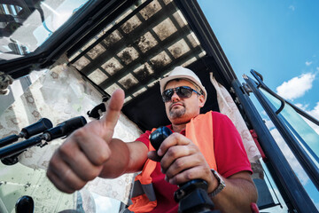 Happy excavator operator in white helmet seated indoor machine cabin showing cool sign on summer day