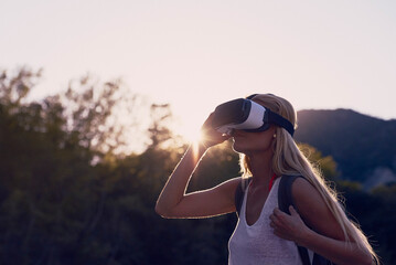 Woman hiking at sunrise using VR goggles to explore the metaverse. Blond woman on holiday hiking in...