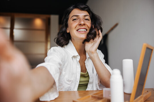 Smiling young caucasian woman with snow-white smile makes photo sitting in room. Brunette with bob haircut is wearing shirt. Lifestyle and beauty concept