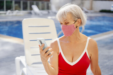 Senior woman in face protective mask ussing smartphone on the sunlounger near the swimming pool.
