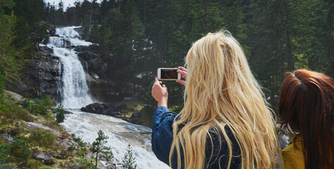 Woman photographing a waterfall on her cellphone during a hike in nature with a friend.Two women on...