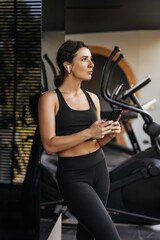 Fototapeta na wymiar Fit young caucasian woman looking away using mobile phone in gym. Brunette wears black sports top and leggings. Healthy lifestyle, technology concept