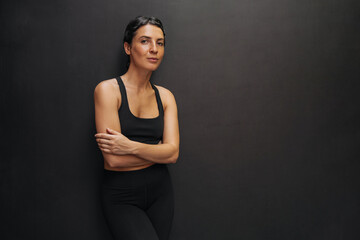 Fototapeta na wymiar Slim young caucasian woman in tracksuit looks at camera on black background with space for text. Brunette is resting after workout. Emotional feeling, calm and peaceful, concept