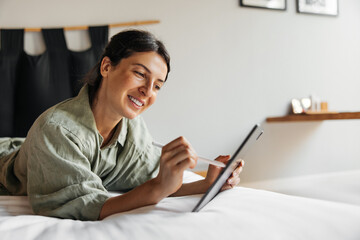 Pretty young caucasian woman uses touchpad of tablet, lies on bed in room. Brunette holds stylus...