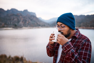 A South Asian man sips coffee from a camping mug with mountains and lake view