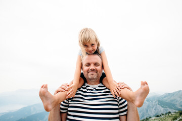 dad and daughter smiling and having fun together. daughter sitting on dad's shoulders and covers dad's eyes with hands. They are standing on the view point on Kotor bay, Montenegro. Wide angle photo