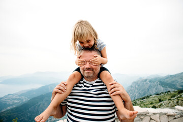 dad and daughter smiling and having fun together. daughter sitting on dad's shoulders and covers dad's eyes with hands. They are standing on the view point on Kotor bay, Montenegro. Wide angle photo