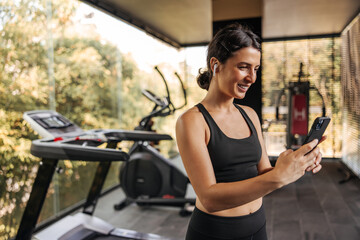 Fototapeta na wymiar Happy young caucasian woman listening to music through headphones from smartphone in gym. Fitness girl wears black top and leggings. Telephone technology concept