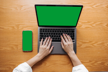 Fototapeta premium Young caucasian woman work at office desk using laptop and smartphone. Blank green screen of wireless devices. Technology concept