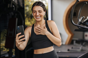 Fototapeta premium Happy young caucasian woman smiling video call via phone in sport club. Brunette wears black top and leggings in training. Technology concept
