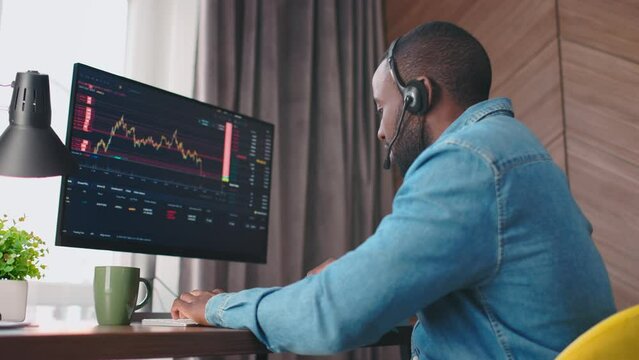 Back side view shot of african american trader in headphones with microphone typing, looking on sceen and speaking. Crypto currency trader. Graphs analysis, stock market. 4k. Office atmosphere.