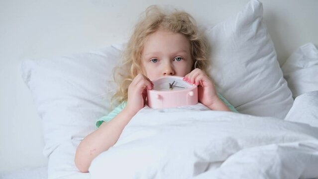 A little sad girl lying on pillow with alarm clock on a white bed and covered with a blanket. early ascents of children to school and kindergarten. comfortable bedding.