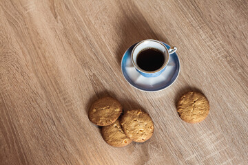 A cup of hot fragrant black coffee in a saucer with cookies, on a wooden light brown textured table.