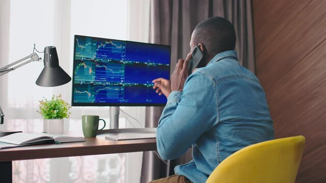 Back view portrait on african american man looking at crypto currency graphs and talking on phone. Crypto currency trader. Graphs analysis, stock market. 4k. Office atmosphere.
